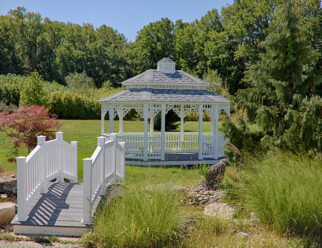 12'x16' Oval White Vinyl Gazebo With Black Shingled Pagoda Roof, Screens, and Cupola In A Backyard With Garden Bridge