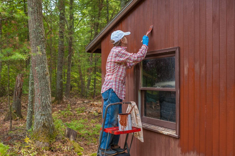 A middle aged woman paints her shed a reddish brown.