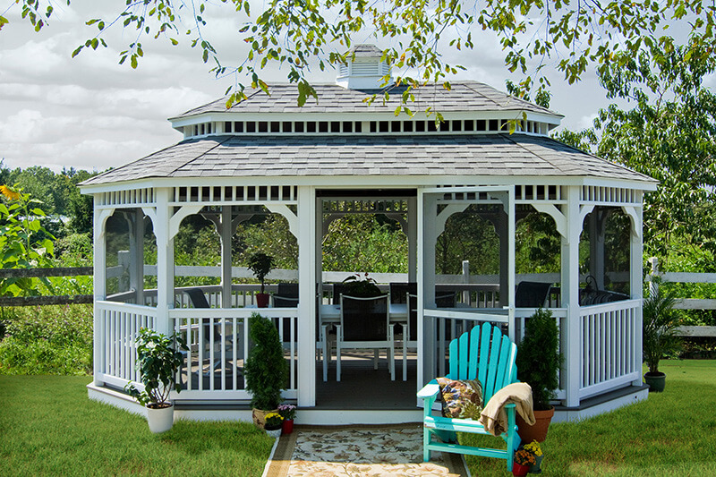 A white, screened-in gazebo is decorated with accent shrubs and has a table set and comfortable chairs inside.