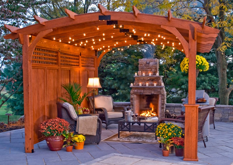 A picture of a wooden pergola on a backyard patio with a fireplace, furniture, and flowers surrounding it.