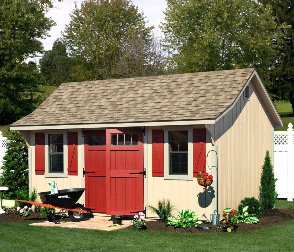 A tan shed with red doors and shutters sits in the corner of a backyard in front of a white fence. Out front is a wheelbarrow and several plants growing in mulch.
