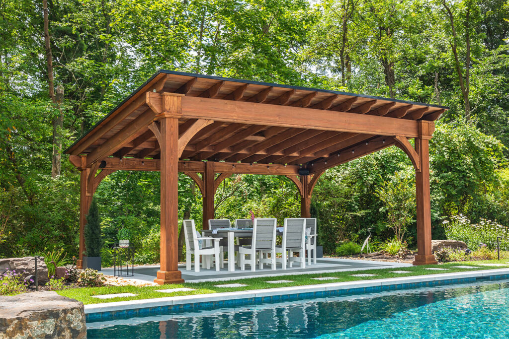 Wooden Santa Fe style pavilion at the side of a pool with outdoor seating beneath it.